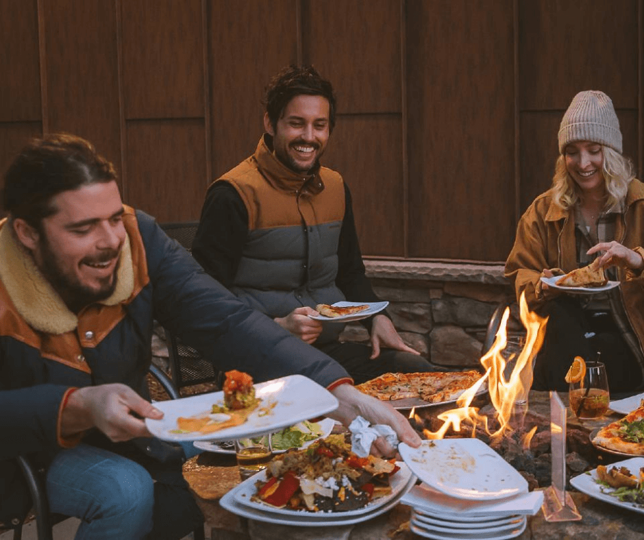 Three people eating food around a gas fireplace.